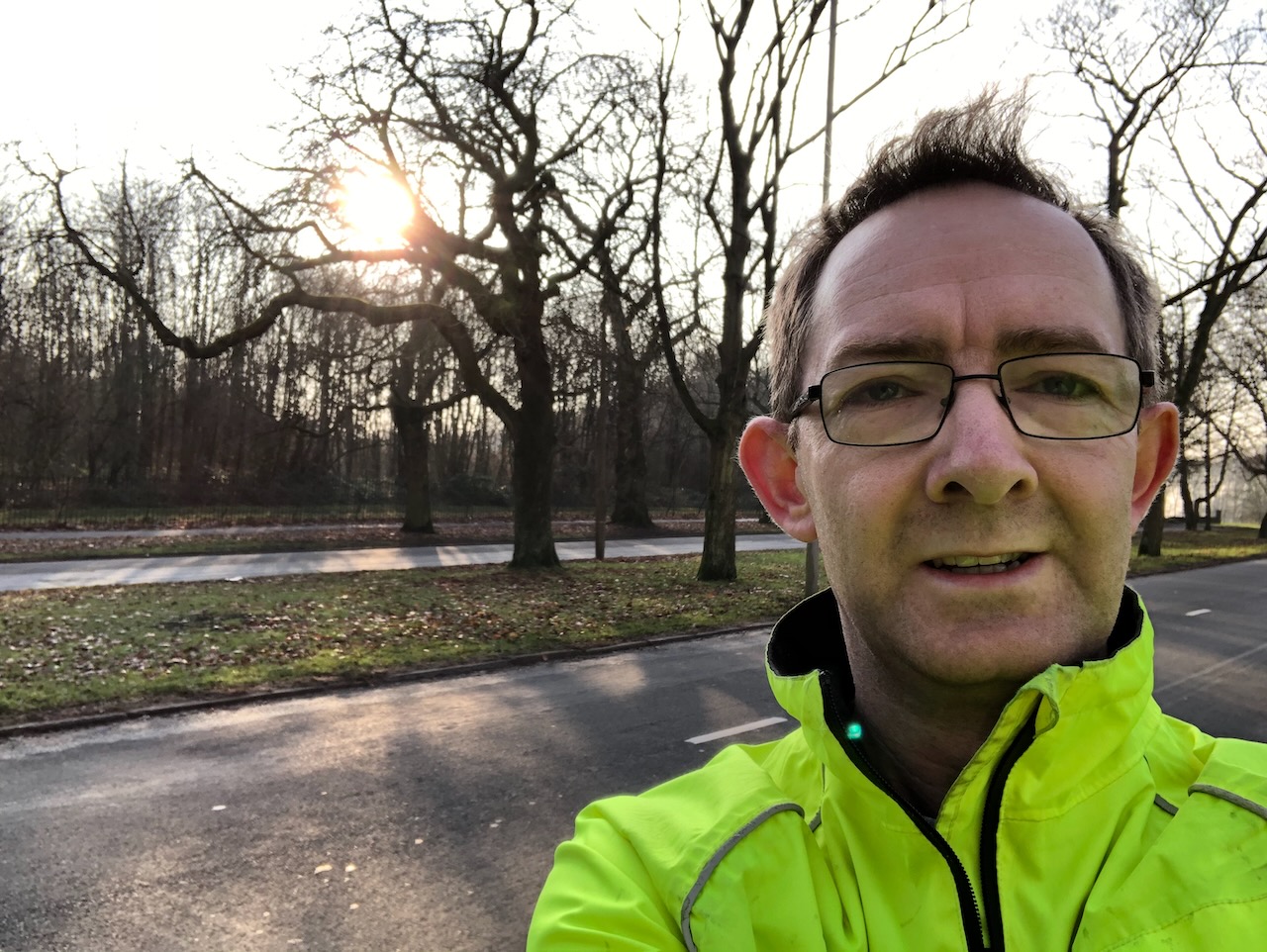 John McKerrell during a run, his head and shoulders are in the foreground wearing a "day glo" green coat, behind is a dual carriageway and bare trees suggesting a winter's day